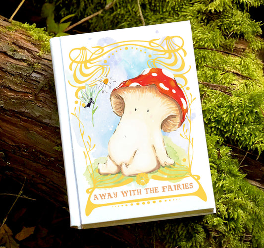 Image shows a white hard backed notebook with a toadstool mushroom seated looking up at a fairy handing her a daisy on the cover. The art has a gold ornate border around it, with a blue background. The text on the front reads "Away with the fairies"