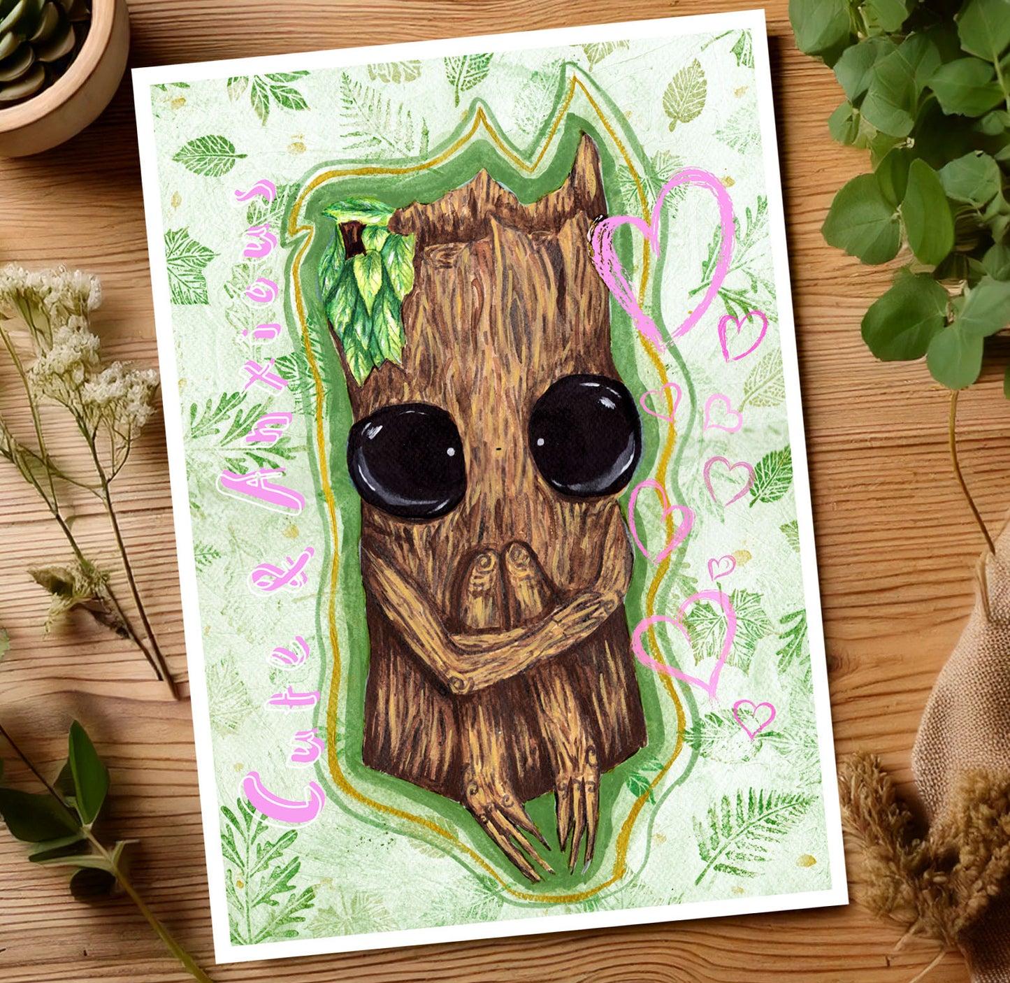 Image features an art print laying on a wooden table surrounded by plants & flowers. The art print features cute and anxious Splint the Twigling, a wooden fantasy creature who is hugging their knees in a seated position. They have big black eyes & are painted onto a green background with green leaves surrounding them. The pink words read, 'Cute and Anxious' to the left of Splint, & to the right of them are a selection of pink hearts in various sizes. Splint is surrounded by a green & golden glow