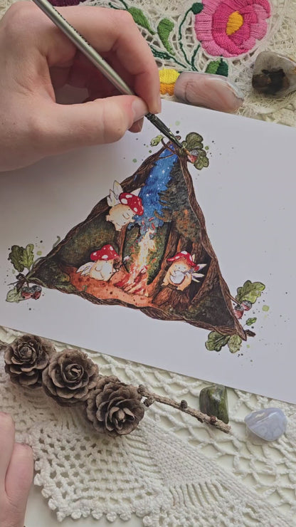 video of artist Matilda Moon adding embellishments of glitter to high quality art print showing three toadstool mushroom fairies around a glowing campfire within a dark forest

the scene is framed within a triangle of vines and branches with acorns and leaves at each point