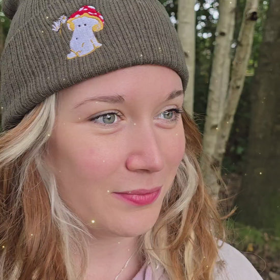 model wears a khaki green ribbed beanie hat with an embroidered image of a fly agaric toadstool mushroom seated, a fairy is beside the mushroom handing them a daisy

model is filmed in front of trees in a forest as whimsical music plays