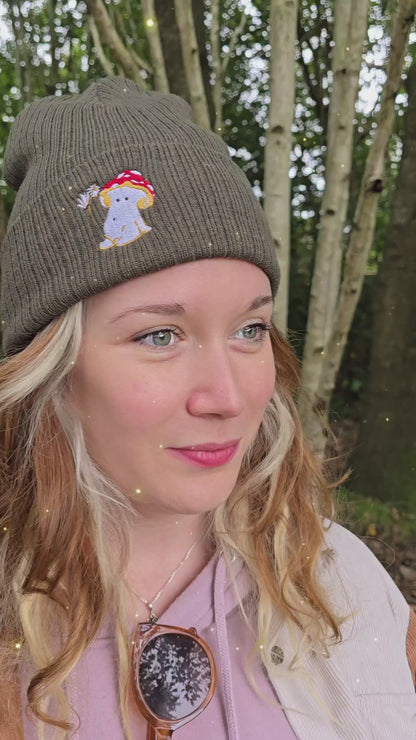 model wears a khaki green ribbed beanie hat with an embroidered image of a fly agaric toadstool mushroom seated, a fairy is beside the mushroom handing them a daisy

model is filmed in front of trees in a forest as whimsical music plays