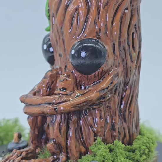 a short video showing hand made sculpture of Splint the Twigling, a tree stump with big black eyes and an anxious expression hugs their knees as they are seated upon a wooden disc, surrounded by moss, grass, rocks and a tiny snail friend. Splint is slowly spinning around 360 degrees so you can see their details from all sides. Splint has green leaves coming from the top of their head, and is photographed against a white background with leaves and plants behind the sculpture