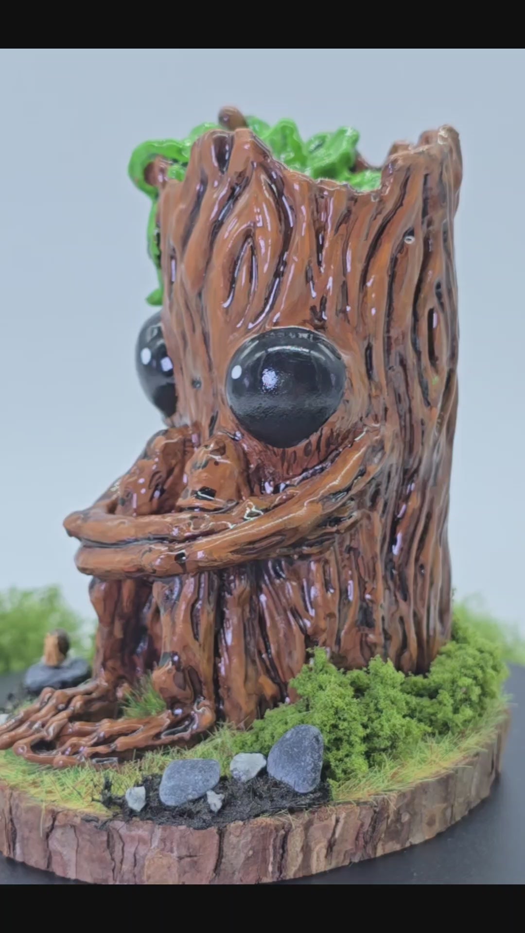 a short video showing hand made sculpture of Splint the Twigling, a tree stump with big black eyes and an anxious expression hugs their knees as they are seated upon a wooden disc, surrounded by moss, grass, rocks and a tiny snail friend. Splint is slowly spinning around 360 degrees so you can see their details from all sides. Splint has green leaves coming from the top of their head, and is photographed against a white background with leaves and plants behind the sculpture