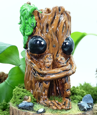 hand made sculpture of Splint the Twigling, a tree stump with big black eyes and an anxious expression hugs their knees as they are seated upon a wooden disc, surrounded by moss, grass, rocks and a tiny snail friend. Splint has green leaves coming from the top of their head, and is photographed against a white background with leaves and plants behind the sculpture