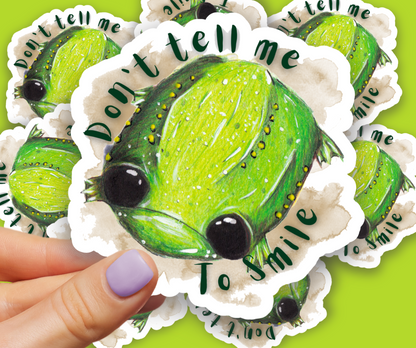 Image shows a sticker placed beside the mousepad of a silver laptop. The sticker features an illustration of Glumbert the Sad  Toad, Glumbert is green with white and yellow spots, he has big black eyes and a sad downturned mouth. Around his body is a brown watercolour smugde and the words, "Don't tell me to smile". The Sticker is held in front of a scattered pile of the same sticker design placed on a bright green background.