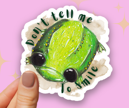 Image shows Glumbert the sad toad, a green spotted toad with a sad mouth and big black eyes. He is placed upon a brown watercolour marking with the words 'Don't tell me to smile' in dark green. The sticker is held between a finger and thumb over a pastel pink background
