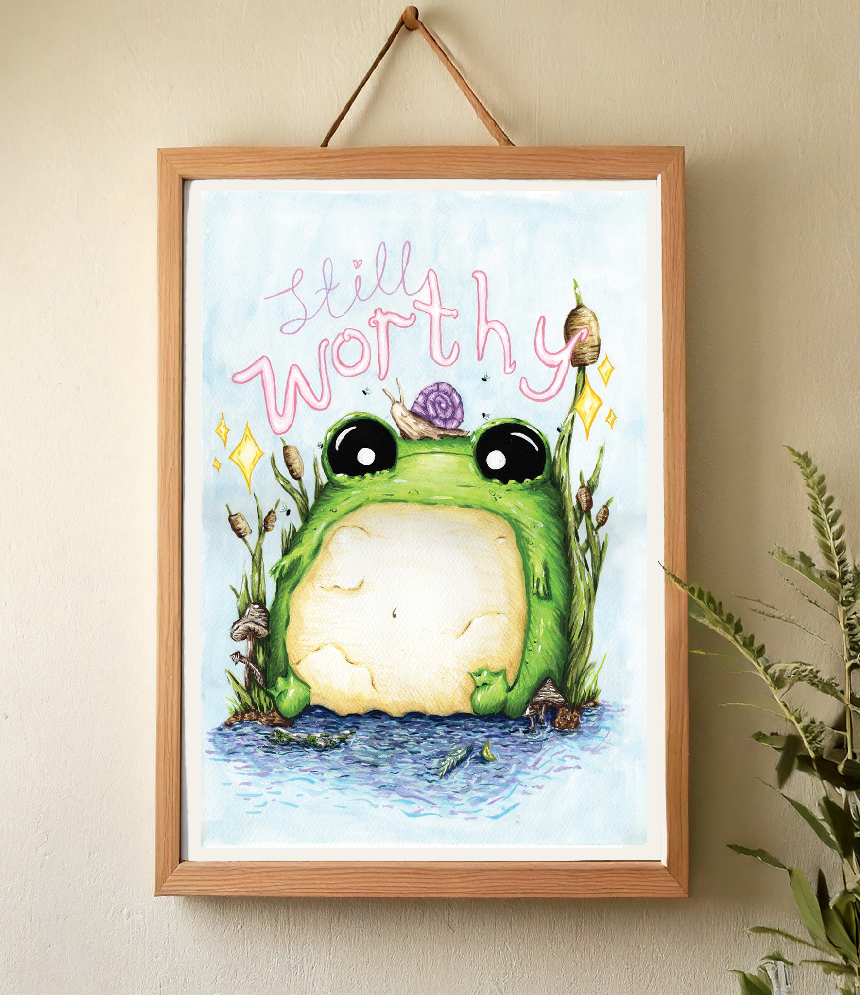 Framed artwork of a green frog / toad with 'Still Worthy' text on a wall.