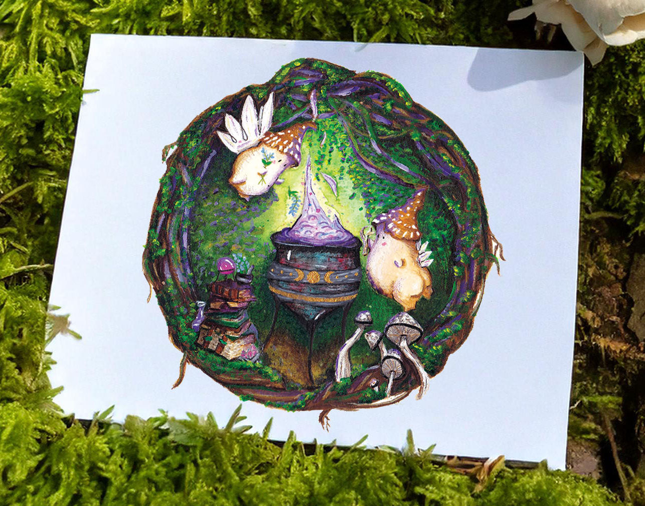 An art print with magical witch mushroom folk around a cauldron brewing a purple glowing potion is resting on a mossy rock. The art print rests on a moss covered tree stump with mushrooms growing from the moss